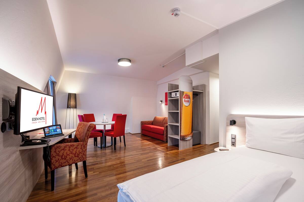 A hotel room with a bed, a desk with a computer, a red sofa, and a dining area with chairs.