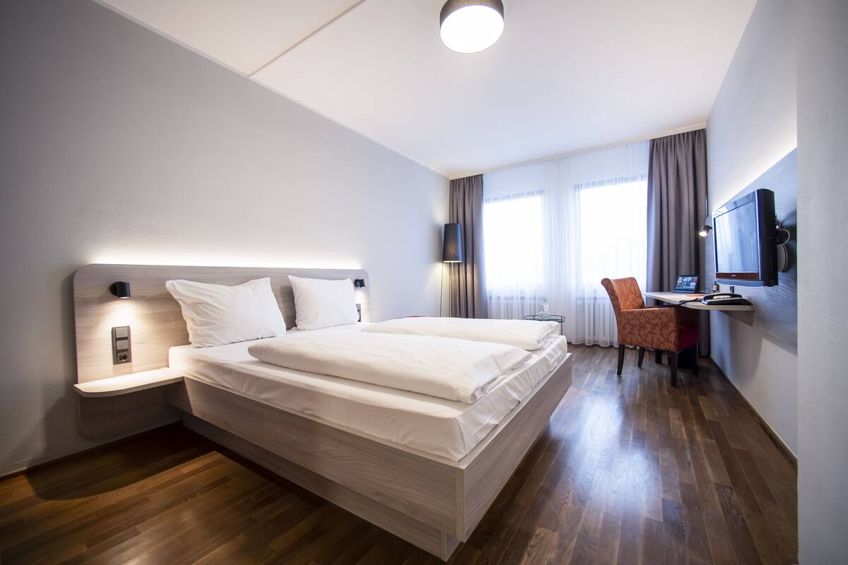 A modern hotel room with a large bed, a desk with a chair, a television, and a window with curtains.