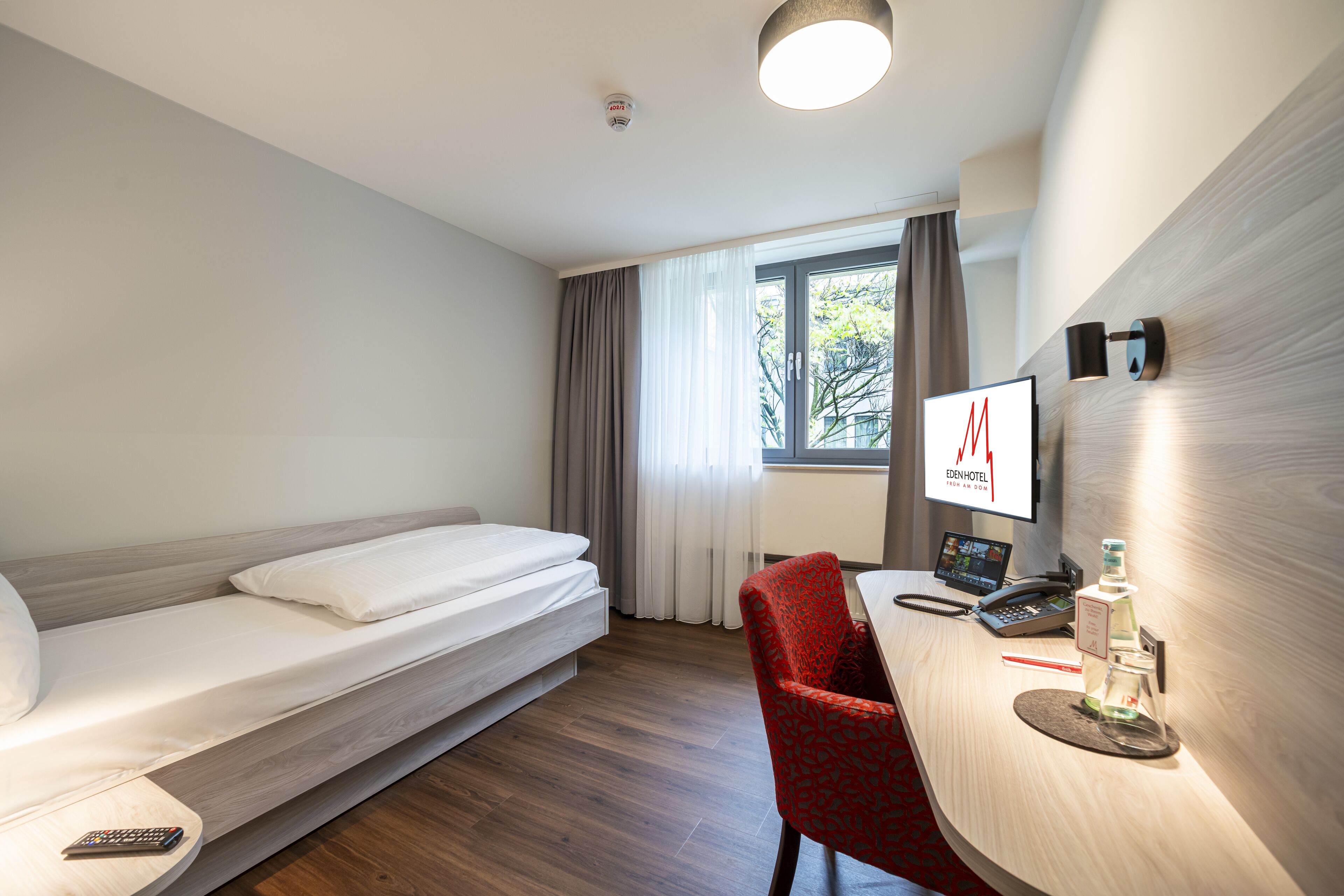 A hotel room with a bed, a desk with a computer, a chair, and a window with curtains.