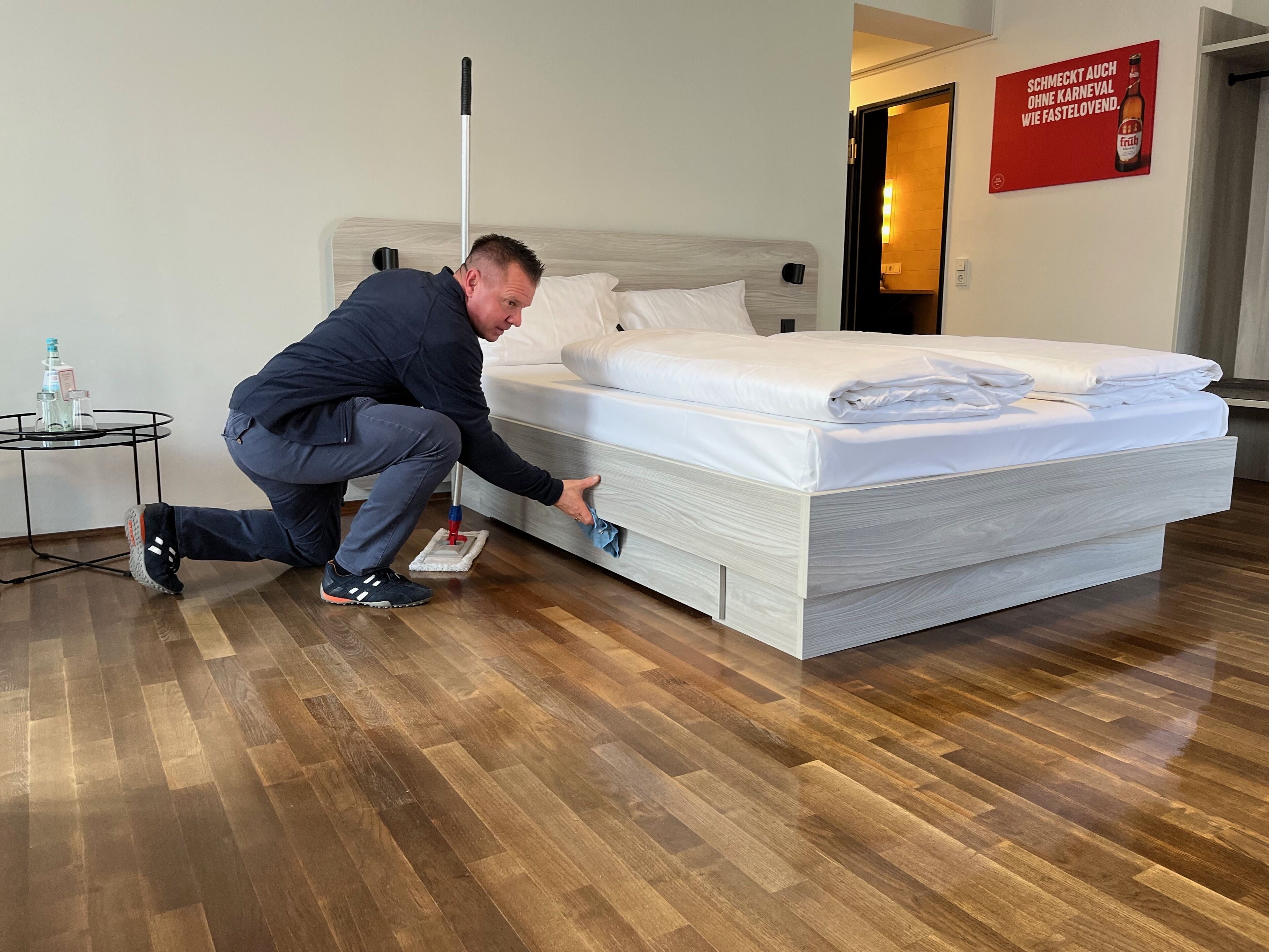 A man cleaning a bed in a hotel room with a wooden floor and a small table with bottles.
