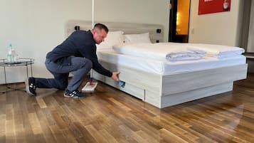 A man cleaning a bed in a hotel room with a wooden floor and a small table with bottles.