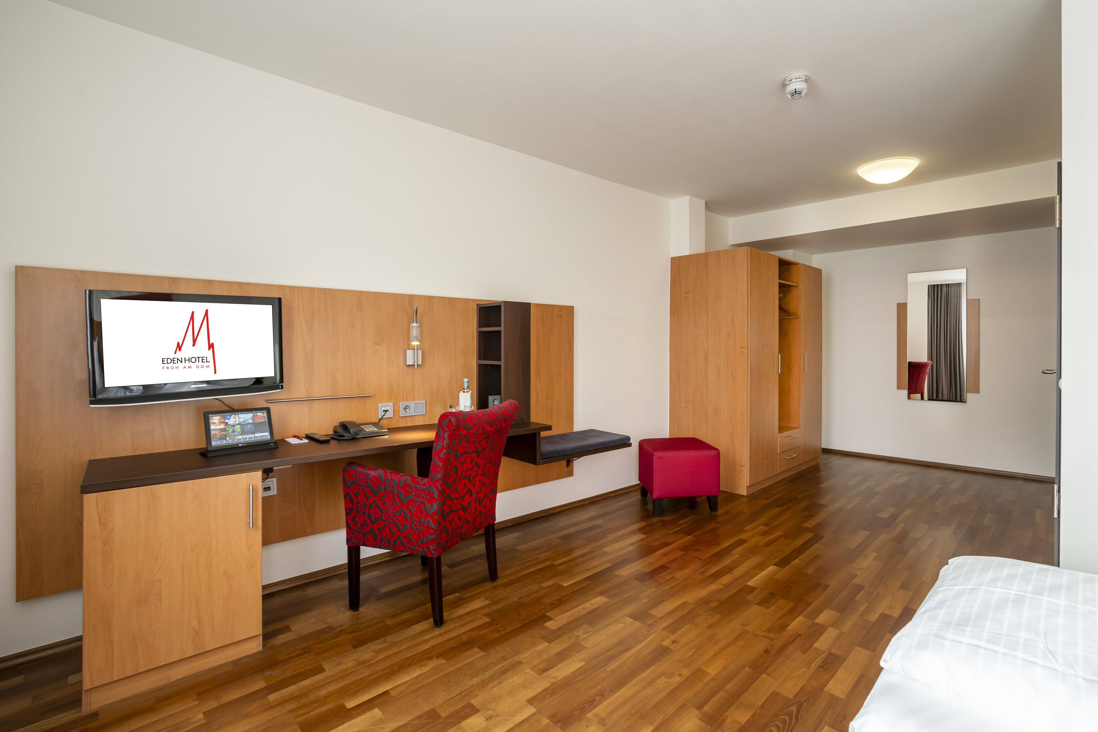 A modern hotel room with a wooden desk, a red patterned chair, a bed, and a TV mounted on the wall.