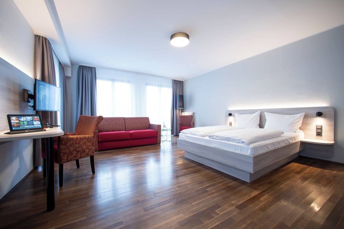 A modern hotel room with a wooden floor, a large bed, a red sofa, a desk with a laptop, and a TV mounted on the wall.