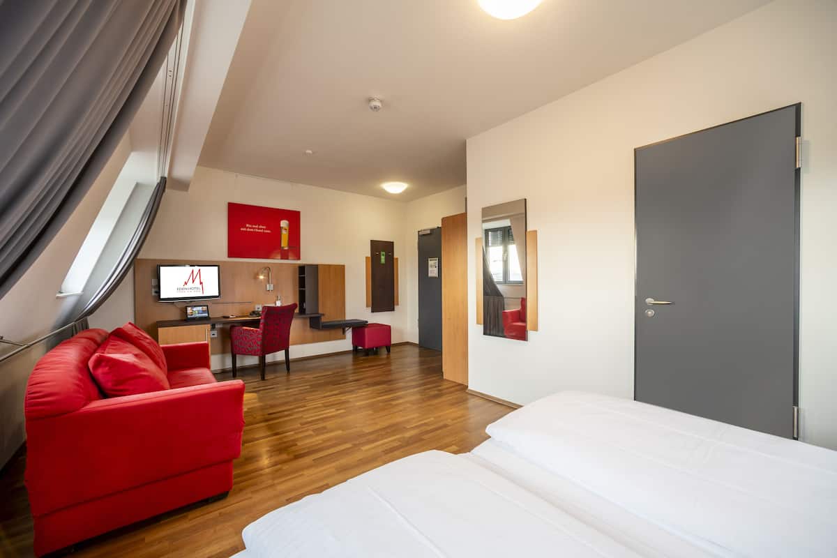 A hotel room with a red sofa, a bed, a desk with a chair, and a TV.
