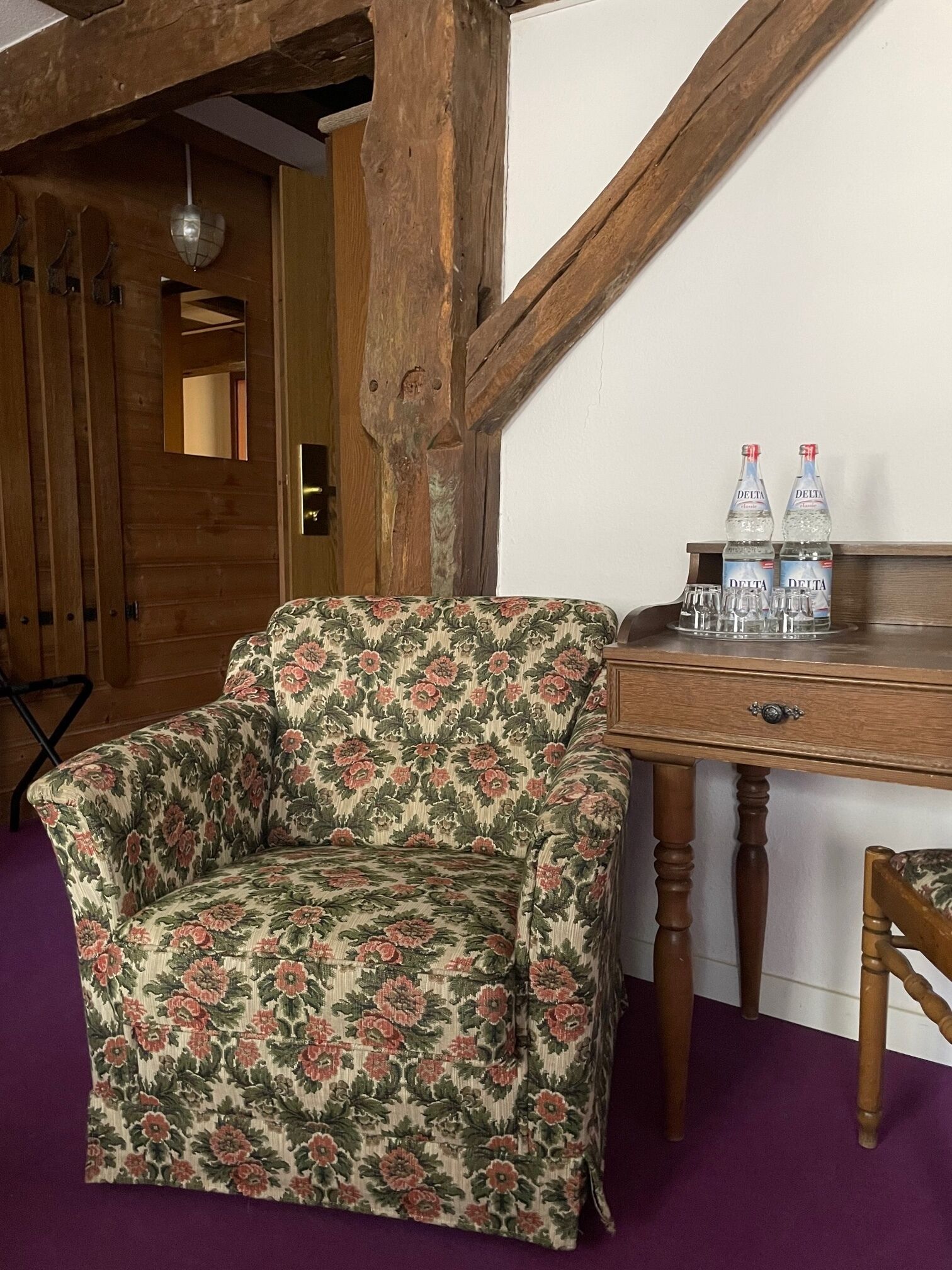 Ein Zimmer mit einem blumengemusterten Sessel, einem Holztisch mit zwei Wasserflaschen und einem Spiegel an der Wand.
