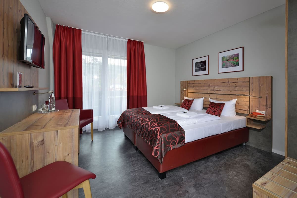 A hotel room with a wooden desk, two red chairs, a bed with white and red bedding, a TV on the wall, and two framed pictures on the wall.