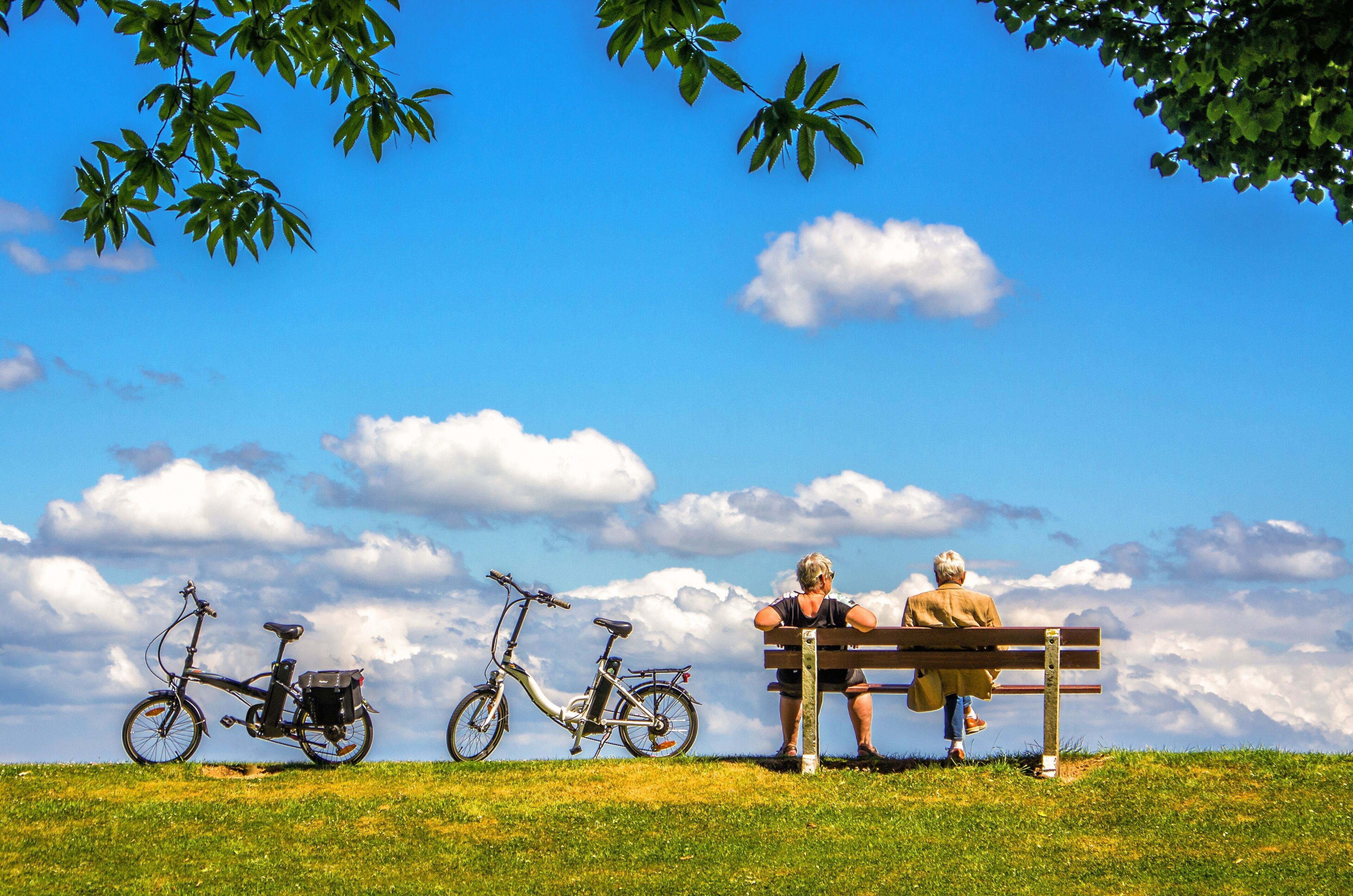 Eine Parkbank mit zwei Personen, einem Fahrrad und blauem Himmel mit Wolken.