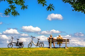 Un banc de parc où sont assis deux personnes, un vélo et un ciel bleu dégagé parsemé de nuages.