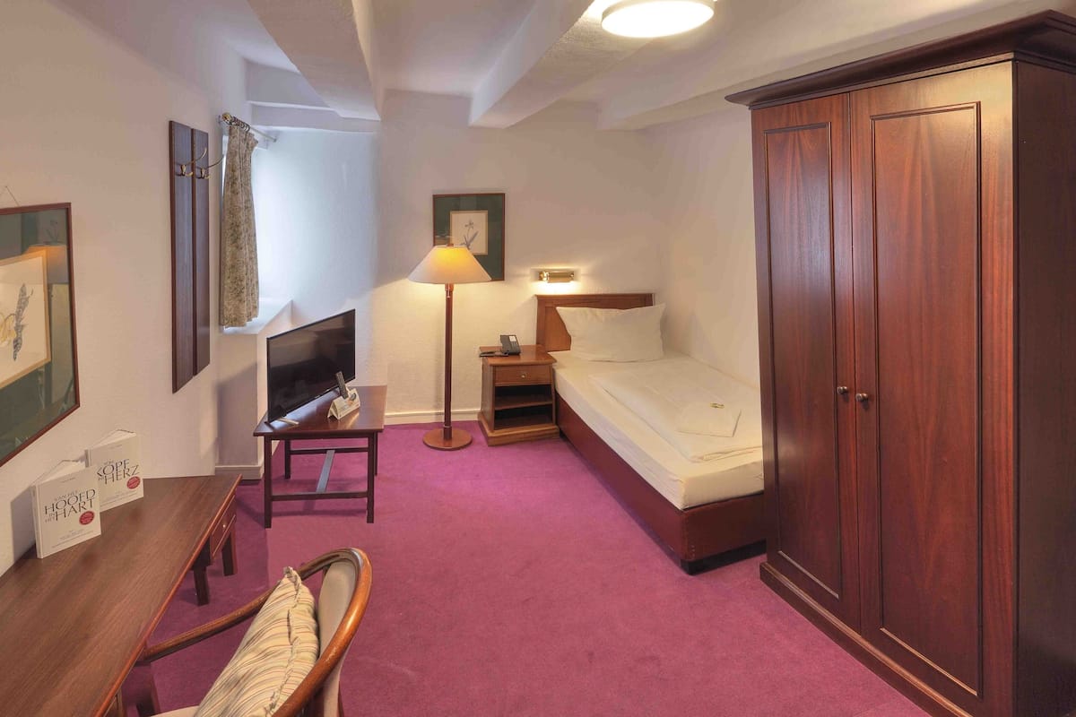 A hotel room with a bed, a desk with a chair, a wardrobe, a TV, and a lamp.