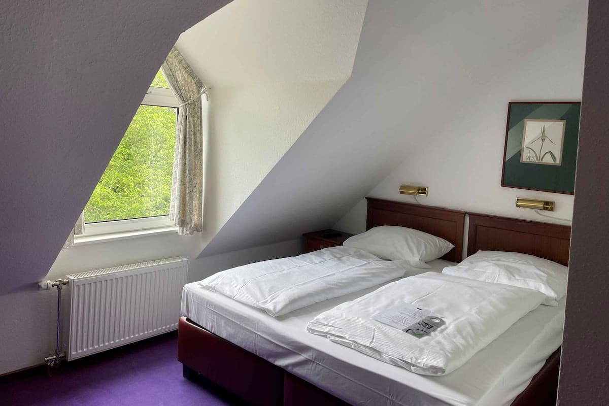 A bedroom with a sloped ceiling, a window with curtains, a radiator, a bed with white bedding, a bedside table, and a framed picture on the wall.