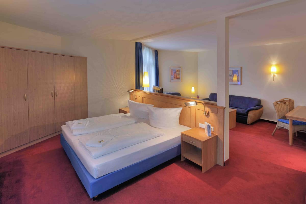 A hotel room with a bed, bedside tables, a wardrobe, a seating area with a chair and table, and wall-mounted lamps.