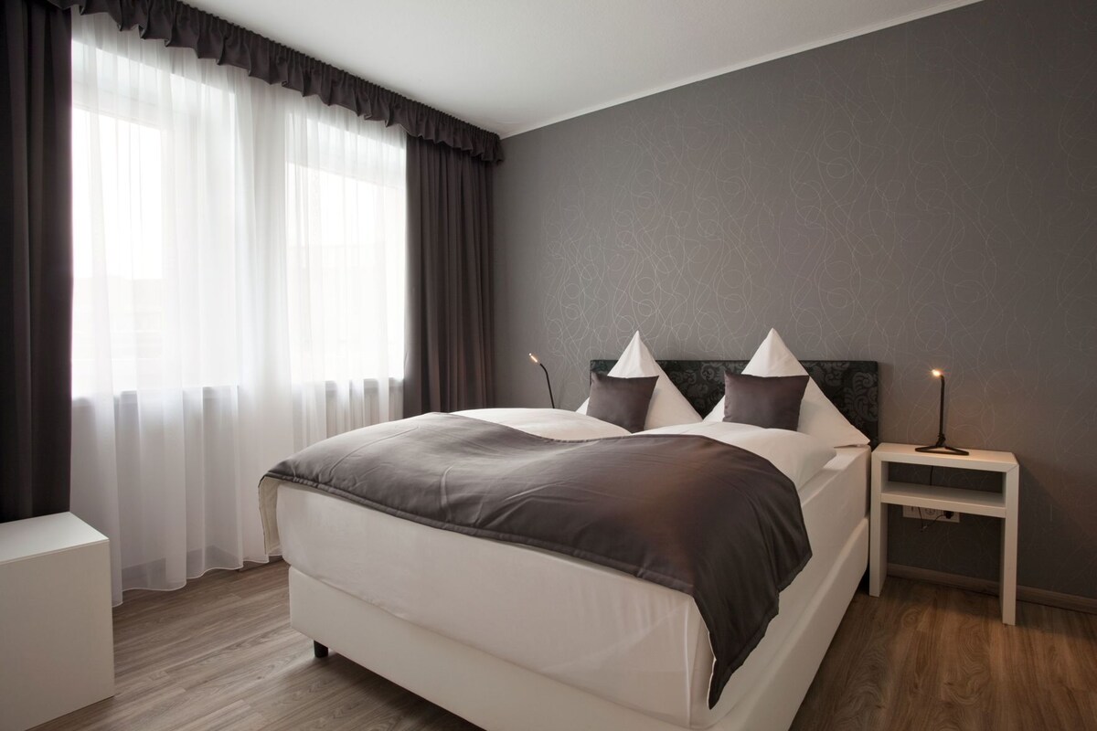 A modern bedroom with a large bed, a bedside table, a nightstand, and a window with sheer curtains.