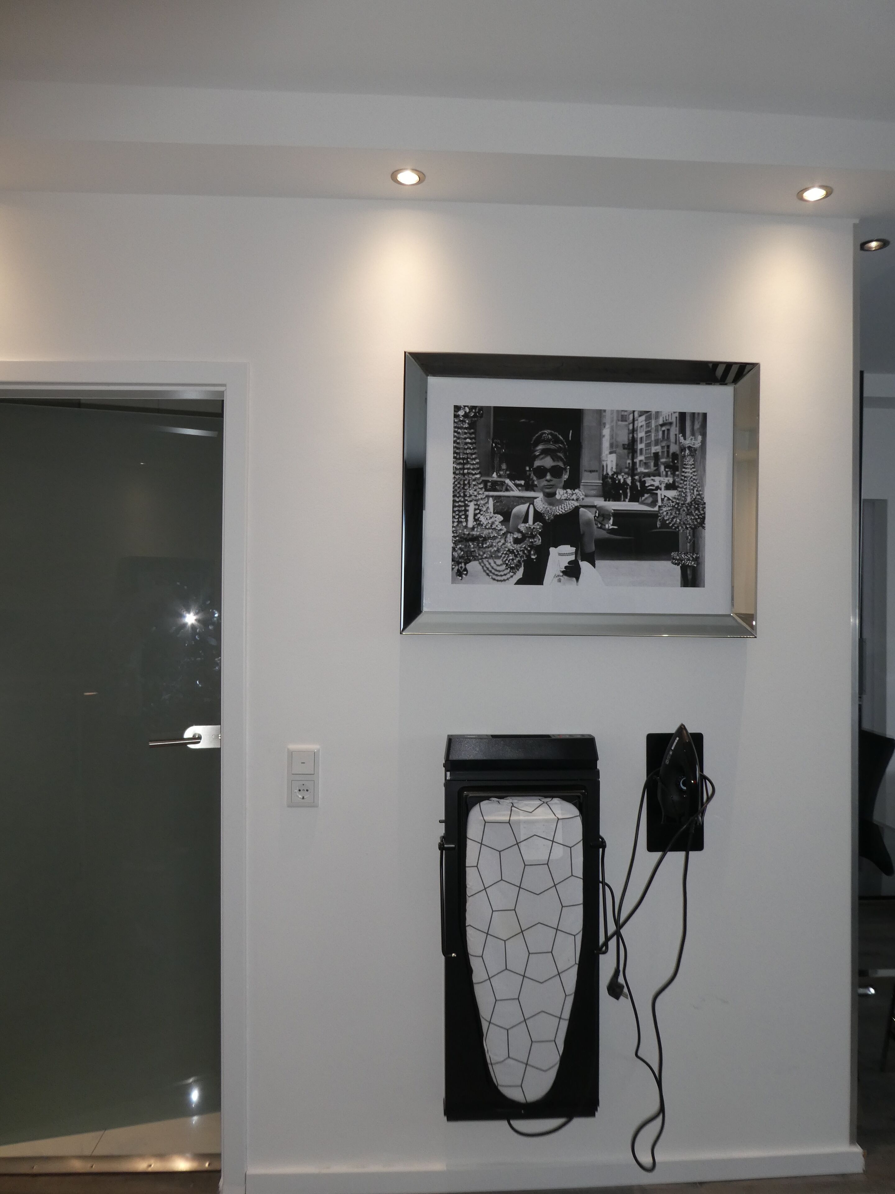 Una parete con una fotografia in bianco e nero incorniciata, un'opera d'arte geometrica e una porta con un pannello di vetro.