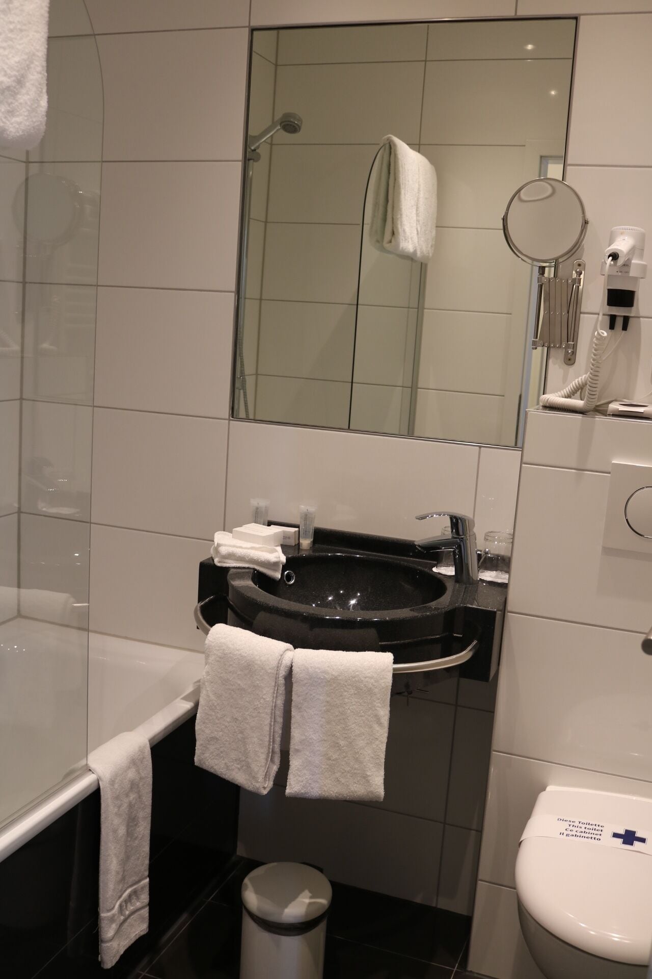 Un baño moderno con ducha envuelta en vidrio, un lavamanos negro con un espejo encima y un inodoro con secador de manos.