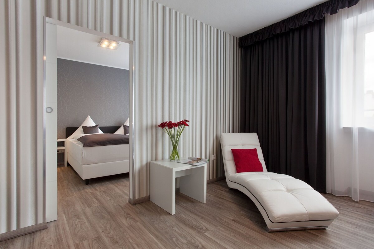 A modern hotel room with a bed, a lounge chair, a small table with a vase of flowers, and a mirror.