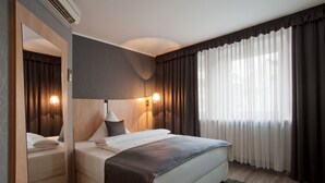 A modern hotel room with a large bed, a bedside table, a mirror, and a window with curtains. - Hotel Asahi (Düsseldorf)