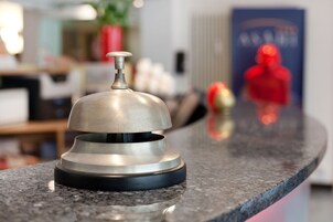 Un oggetto metallico su un piano di lavoro, probabilmente un articolo di servizio alberghiera, con uno sfondo sfocato che include un oggetto rosso e un manifesto.