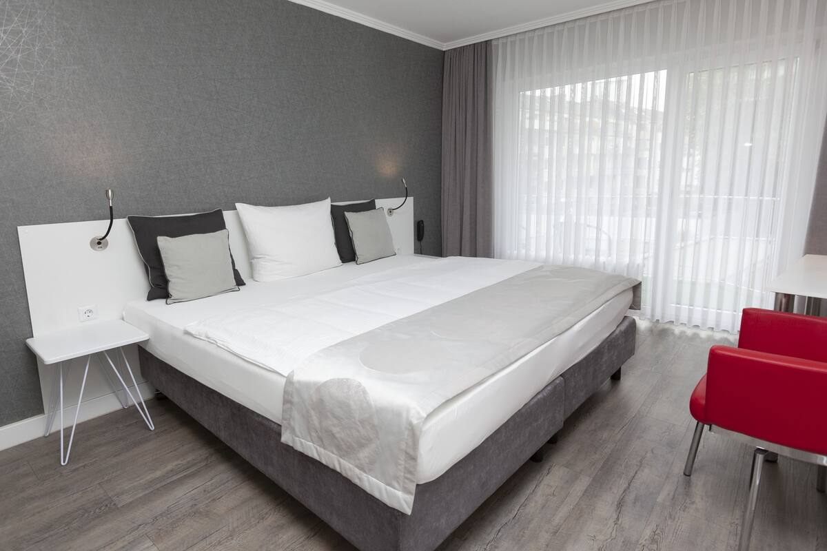 Una habitación de hotel moderna con una cama grande, una mesita de noche, una silla roja y una ventana con cortinas traslúcidas.