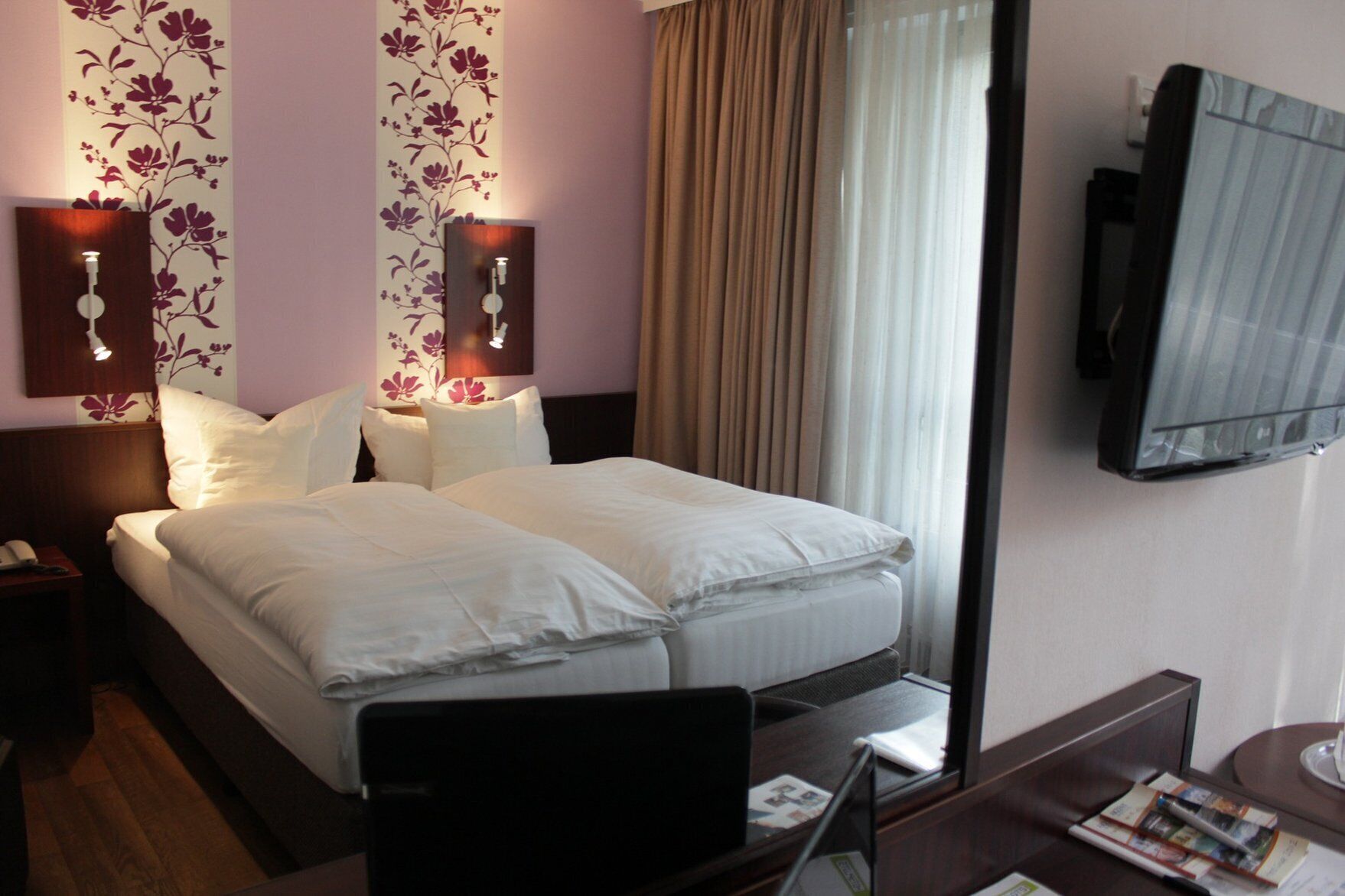 A hotel room with a bed, a desk, a chair, a TV mounted on the wall, and a window with curtains.