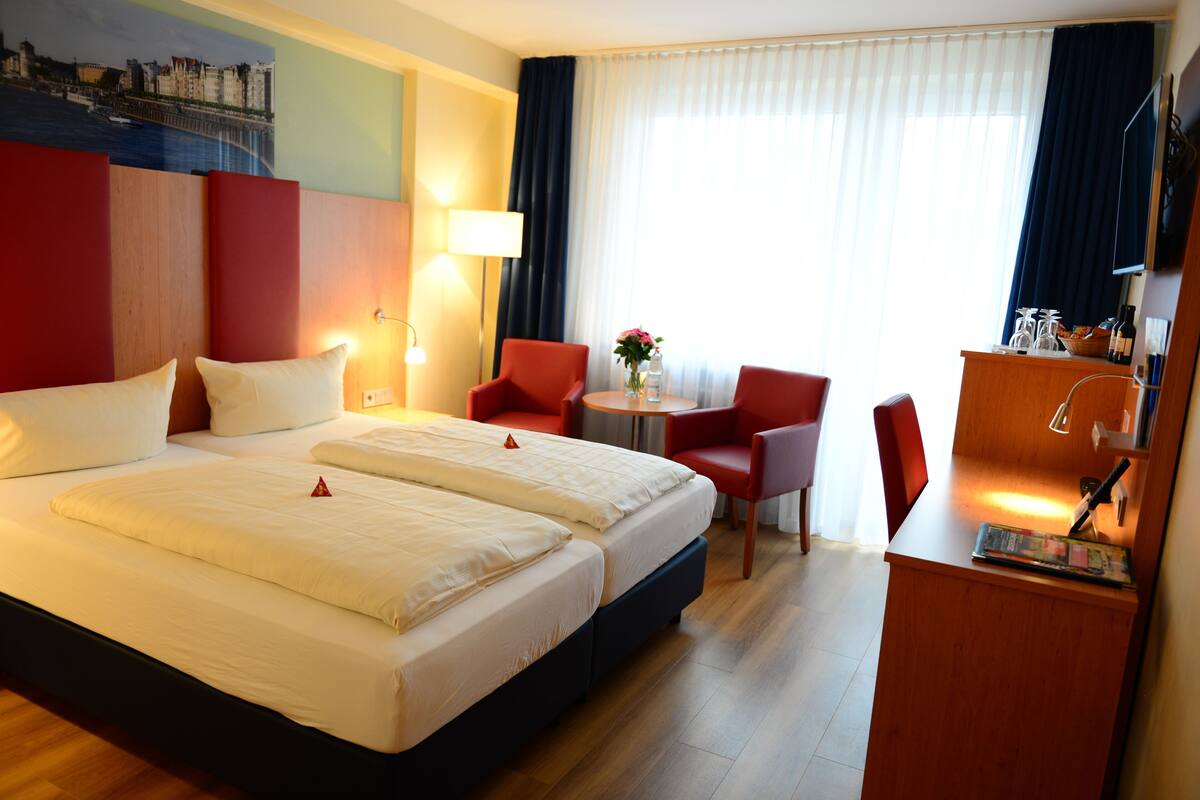A hotel room with a large bed, two red chairs, a small table with a vase, and a desk with a lamp and a bottle.
