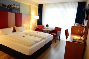 Una camera d'albergo con un letto grande, due poltrone rosse, un tavolino con un vaso e una scrivania con una lampada e una bottiglia.