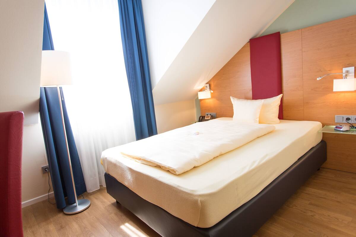 A modern hotel room with a large bed, a bedside table, a lamp, and a view of the outdoors.