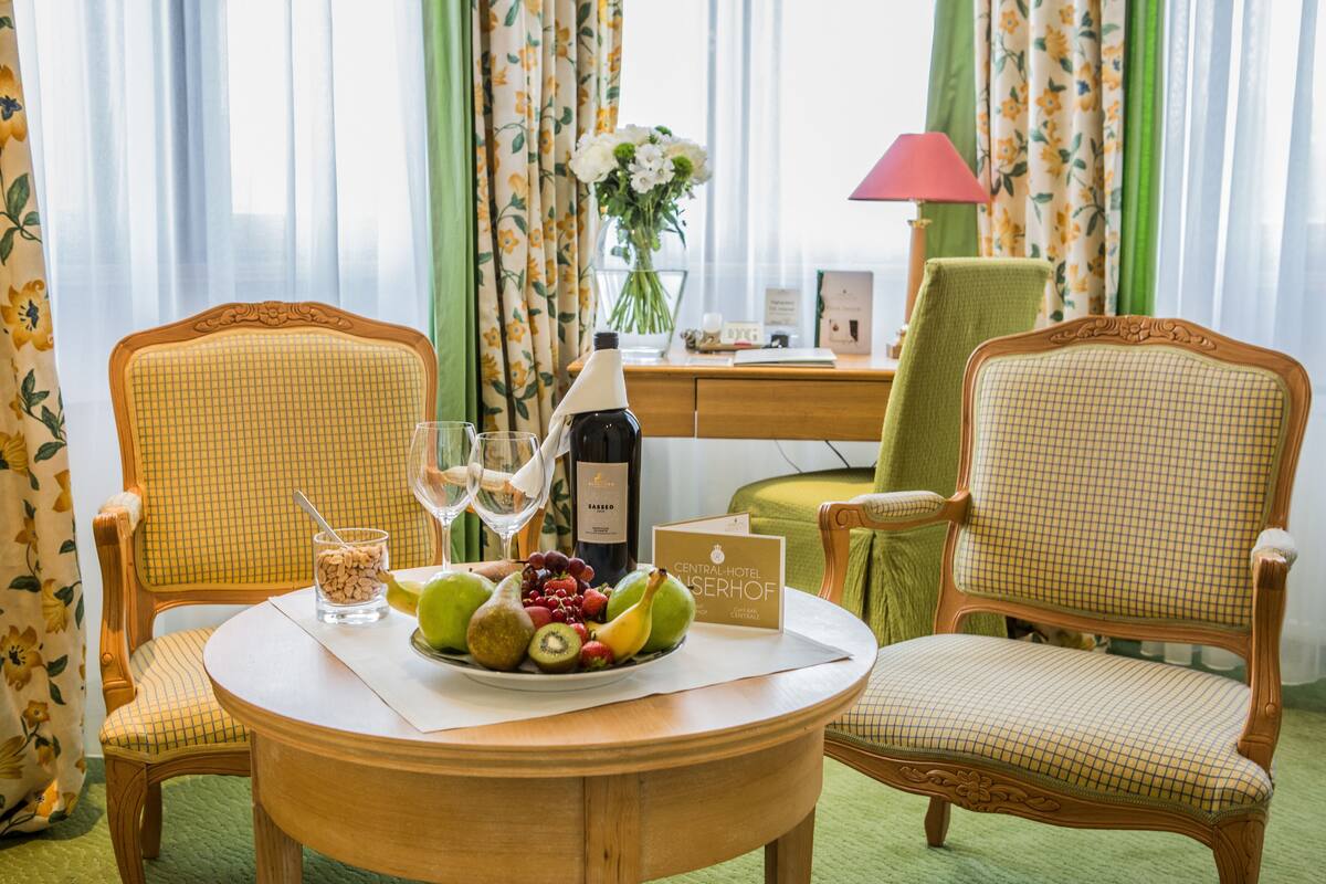 A hotel room with two wooden chairs, a round table with a fruit bowl, wine glasses, and a bottle of wine.