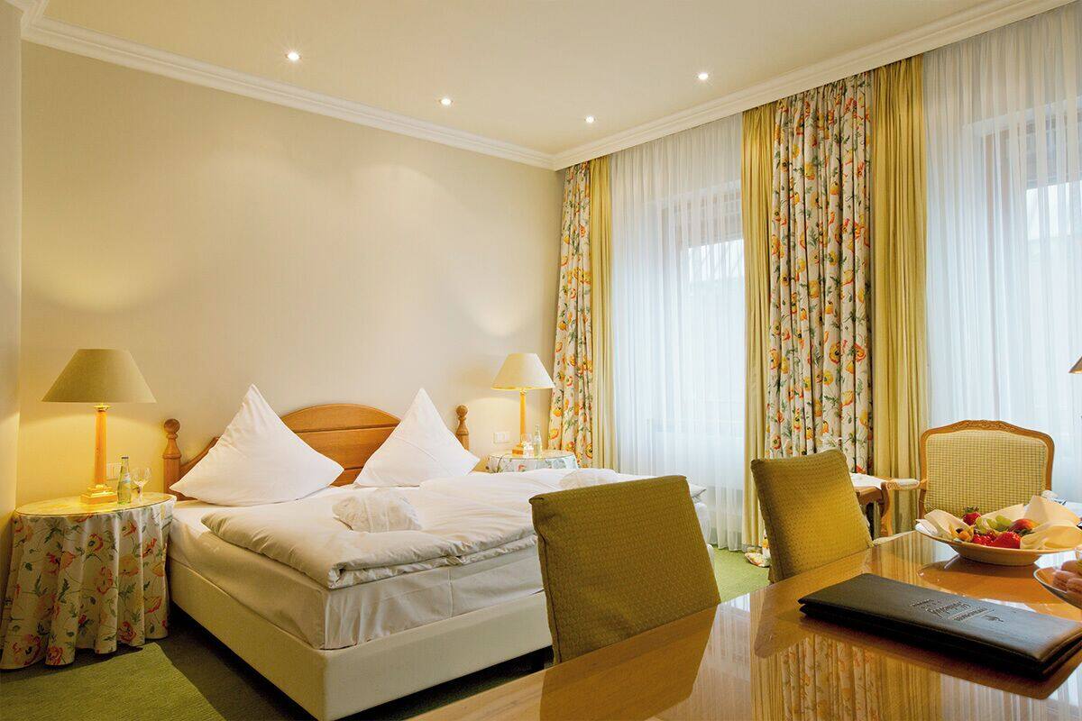 A hotel room with a bed, two bedside lamps, a dining table with chairs, a small table with a lamp, and a fruit bowl.