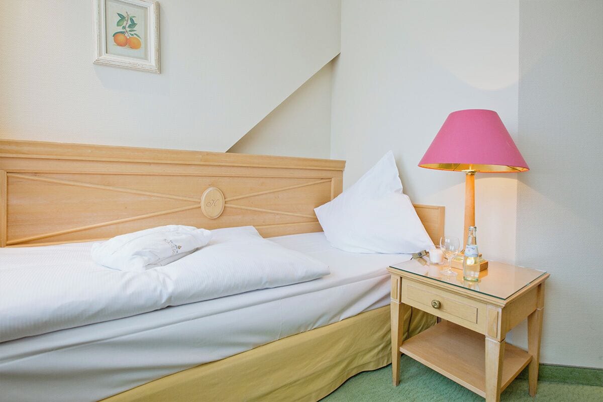 A neatly made bed with white linens and pillows, a wooden headboard, a bedside table with a pink lamp, and a framed picture on the wall.