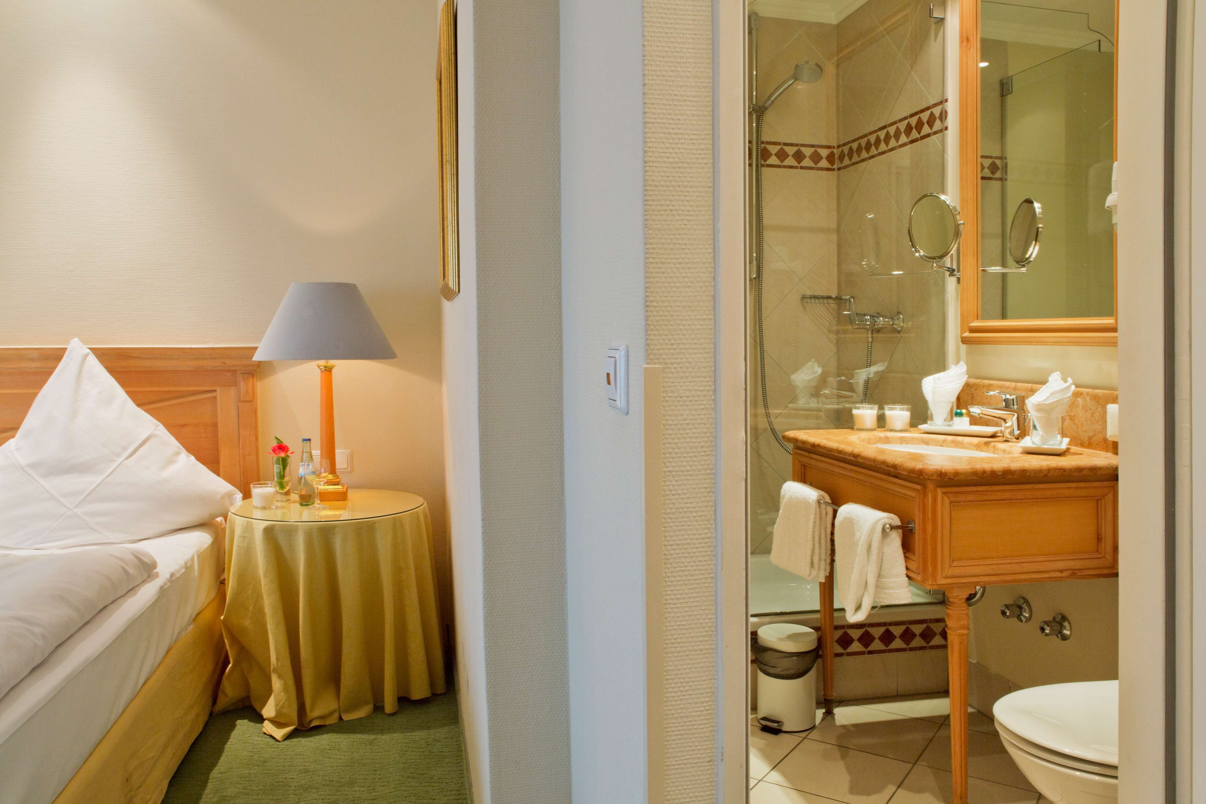 A hotel room with a bed, a bedside table with a lamp, a bathroom with a shower, a sink, and a toilet.