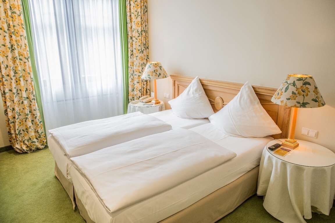 Ein Doppelbett mit weißen Bettwäsche, zwei Nachttische mit Lampen und ein Fenster mit durchsichtigen Vorhängen.