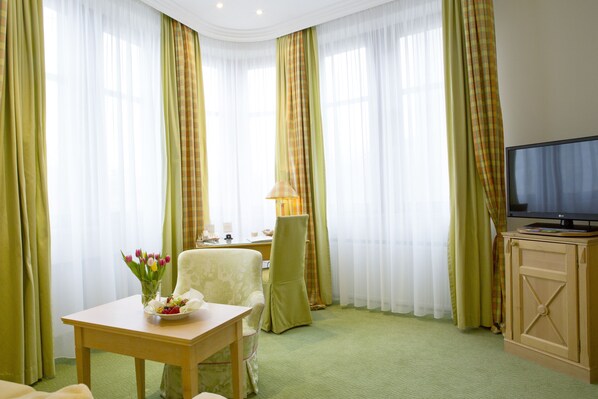 Una habitación de hotel con una mesa de madera, un sillón verde, un televisor sobre un mueble de madera y un escritorio con una lámpara.