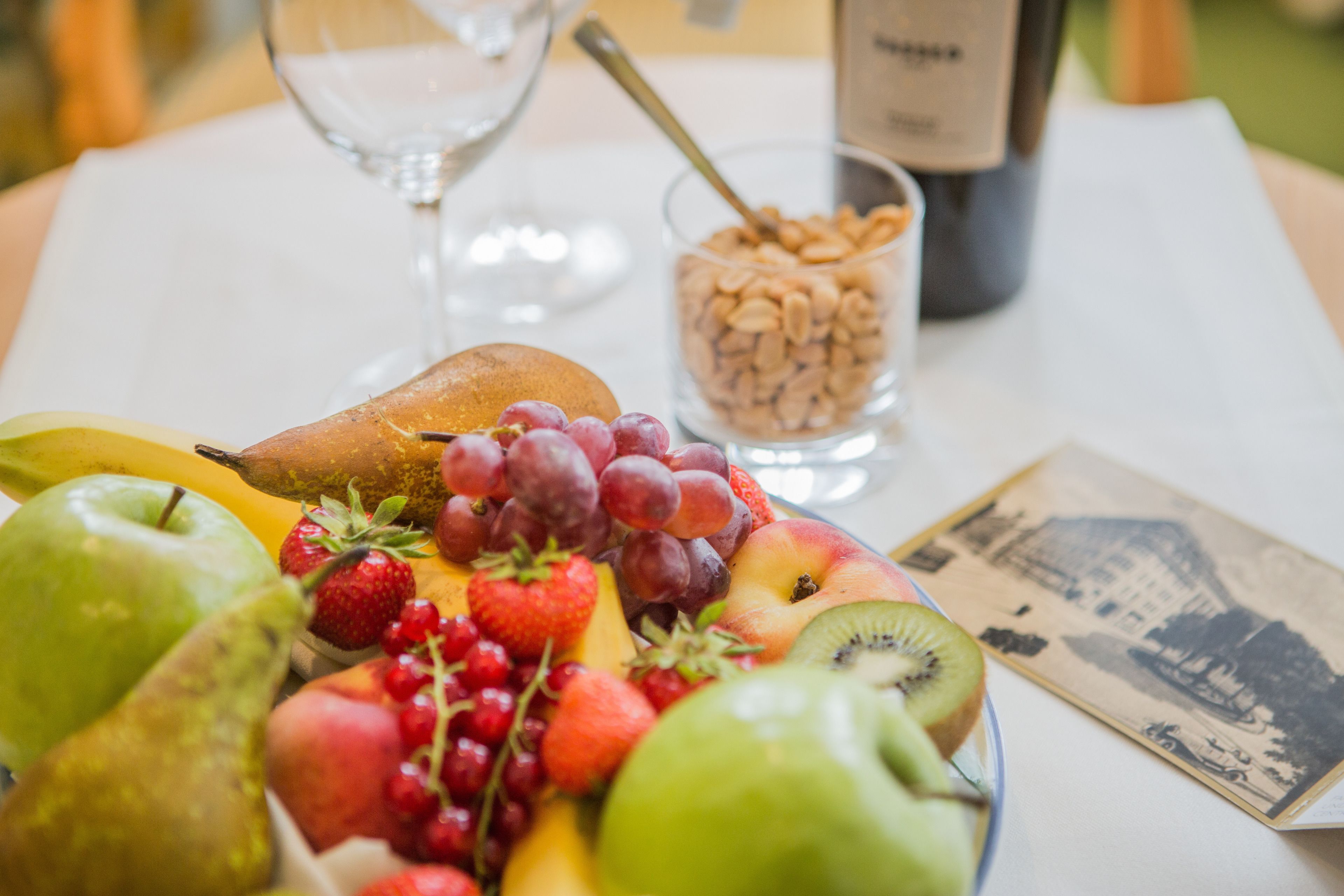 Una mesa con una bandeja de frutas, una botella de vino, una copa de vino y un cuenco pequeño de nueces.