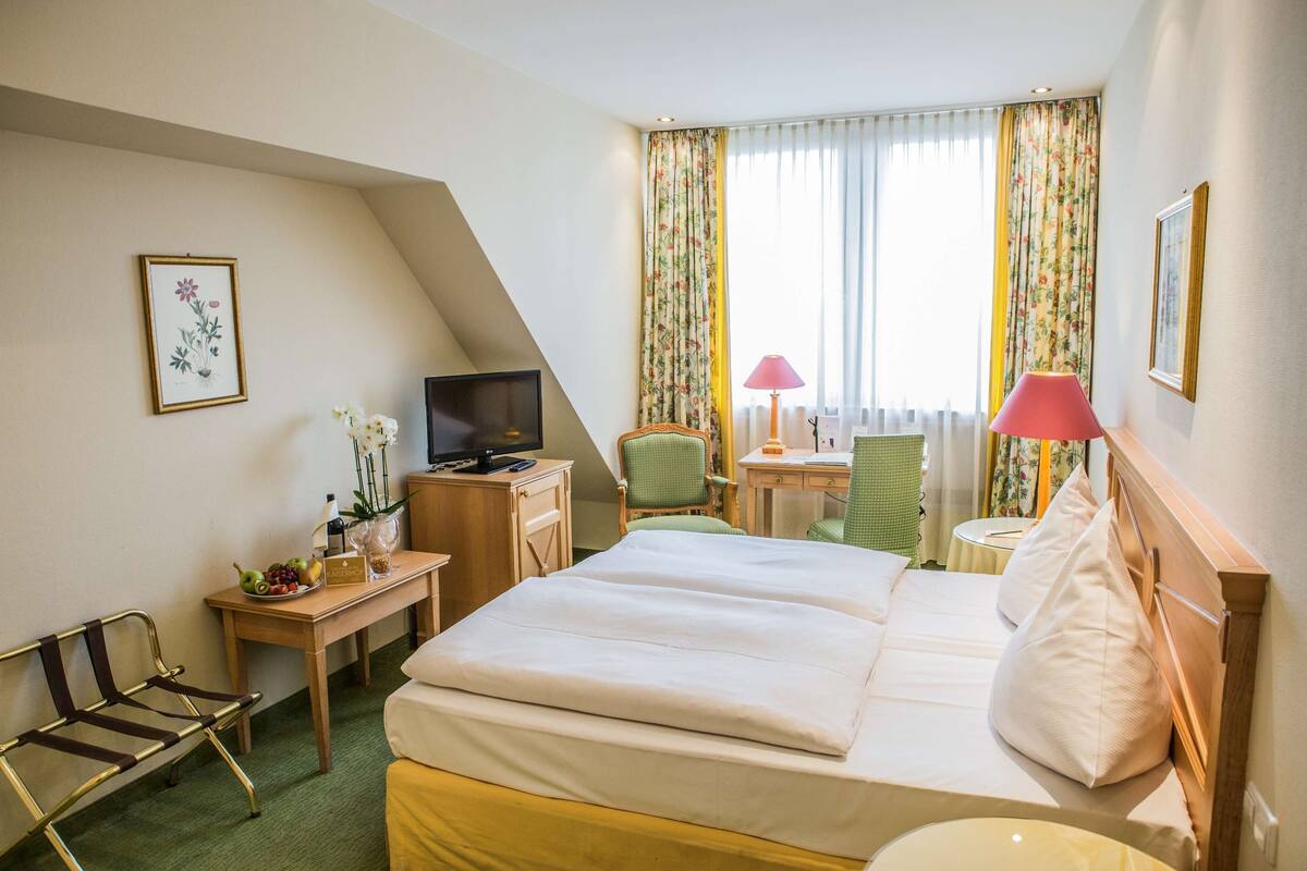 A hotel room with a bed, a desk with a television, a chair, a small table with a lamp, and a window with curtains.