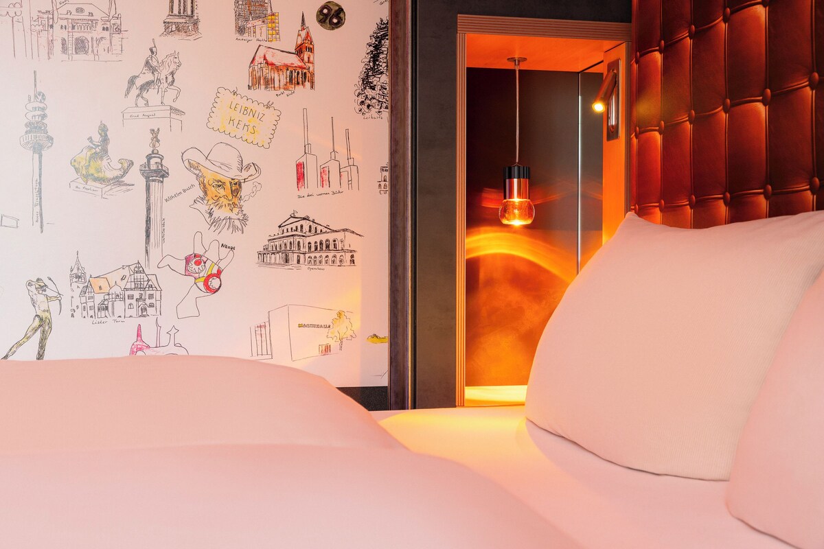 A hotel room with a bed, a wall with various sketches, and a warm light fixture.
