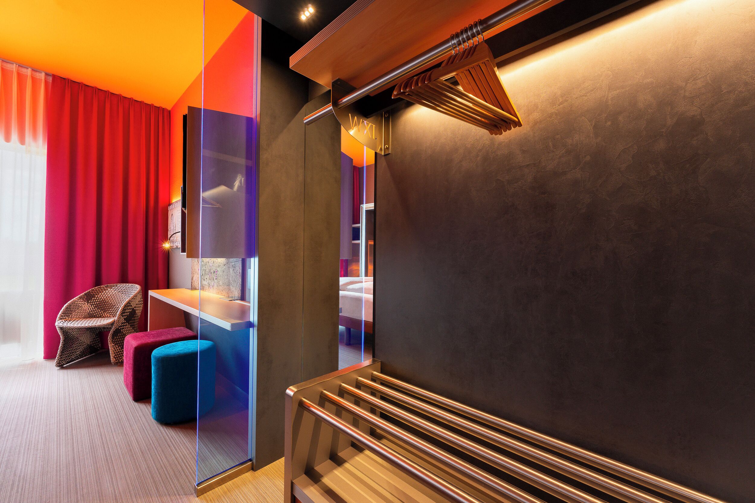 A modern hotel room with a large bed, a seating area with a chair and ottoman, and a glass partition wall.