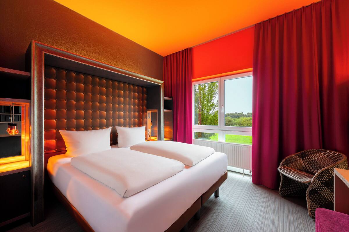 Een moderne hotelkamer met een groot bed, een gestoffeerd hoofdeinde en uitzicht op het omliggende landschap door een raam met rode gordijnen.
