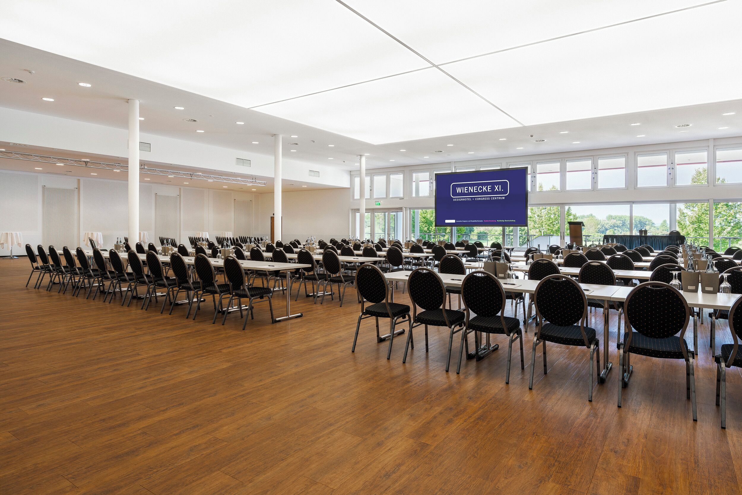 Un amplio salón de conferencias con filas de mesas y sillas, una pantalla grande mostrando 'WIENECKE XL1' y grandes ventanales que ofrecen vistas a zonas verdes exteriores.
