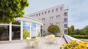 A modern building with a curved glass facade, outdoor seating area with yellow chairs, and a garden with yellow flowers. - Designhotel Wienecke XI. Hannover (Hannover)