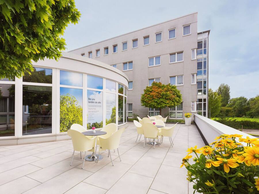 A modern building with a curved glass facade, outdoor seating area with yellow chairs, and a garden with yellow flowers.