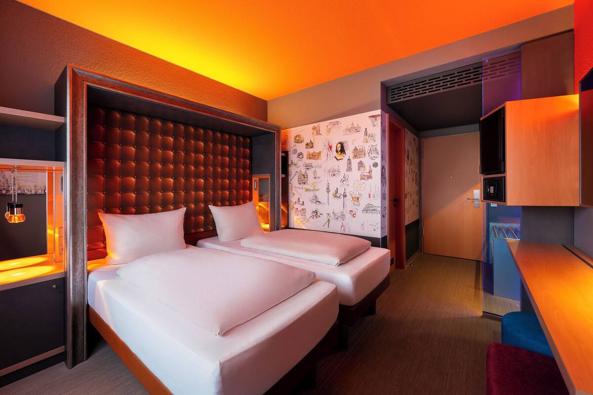 Hotelkamer met twee bedden, een groot gestoffeerd hoofdbord, een muur met illustraties en een klein bureau met lamp.