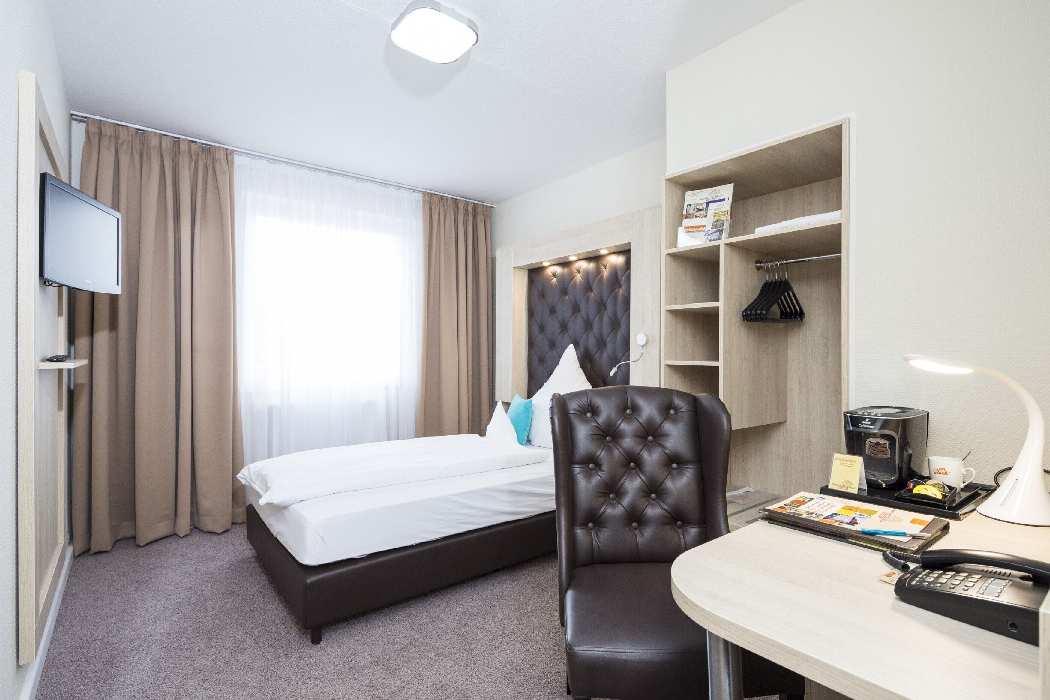Una habitación de hotel con cama, escritorio con lámpara, silla, televisor y armario.