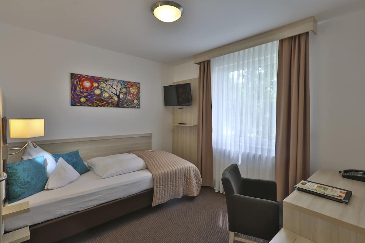 A hotel room with a bed, a desk, a chair, a TV, and a painting on the wall.