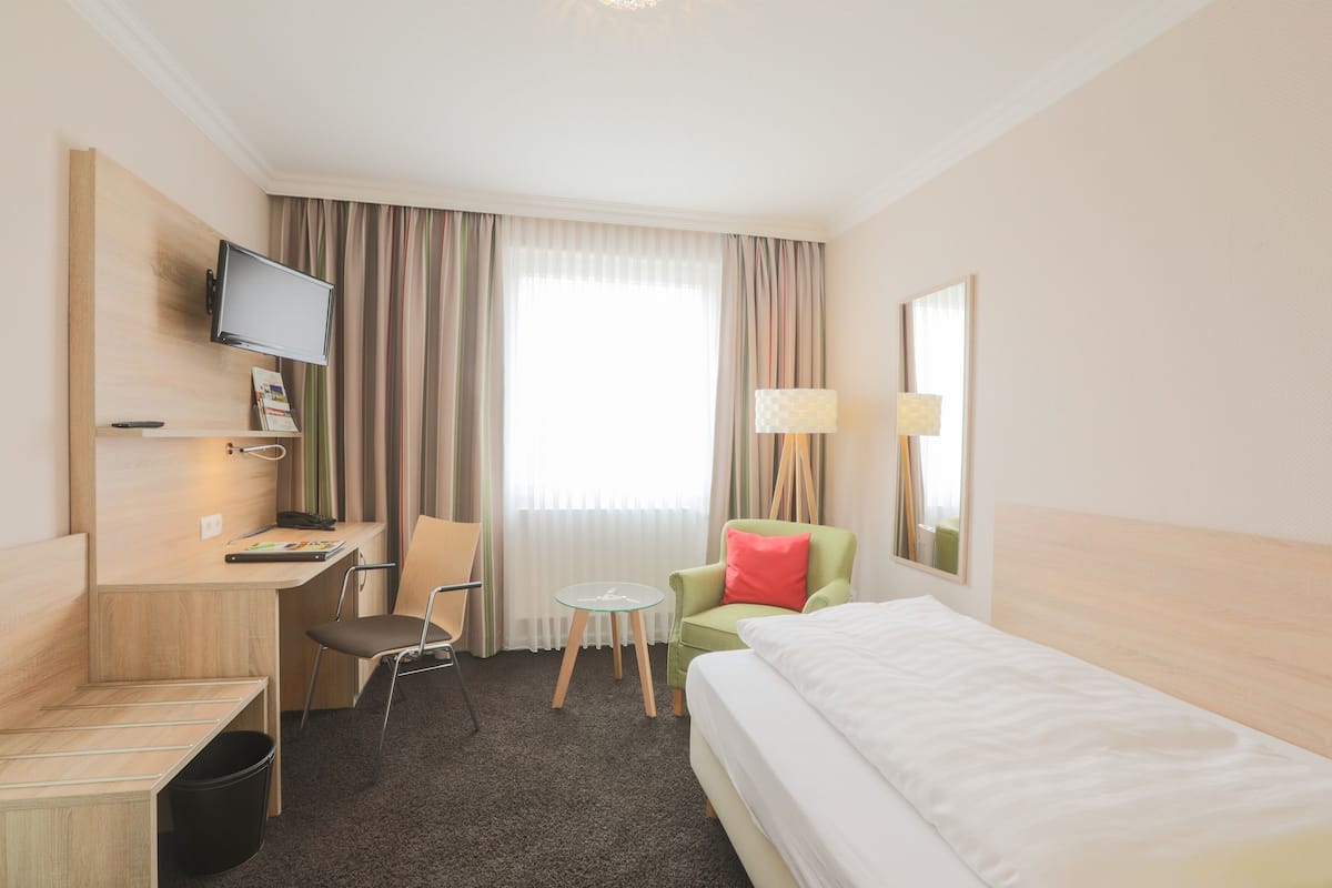 A hotel room with a bed, a desk with a computer, a chair, a small table, and a TV mounted on the wall.