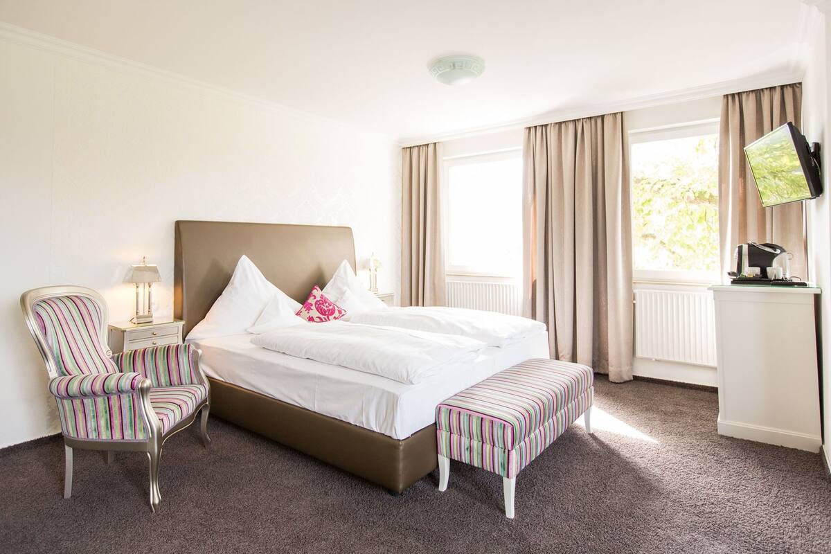 A hotel room with a large bed, a chair, an ottoman, a nightstand, a TV, and a window with curtains.