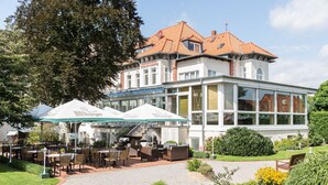 Un hotel con terraza al aire libre, jardín y un edificio grande con tejado de tejas.