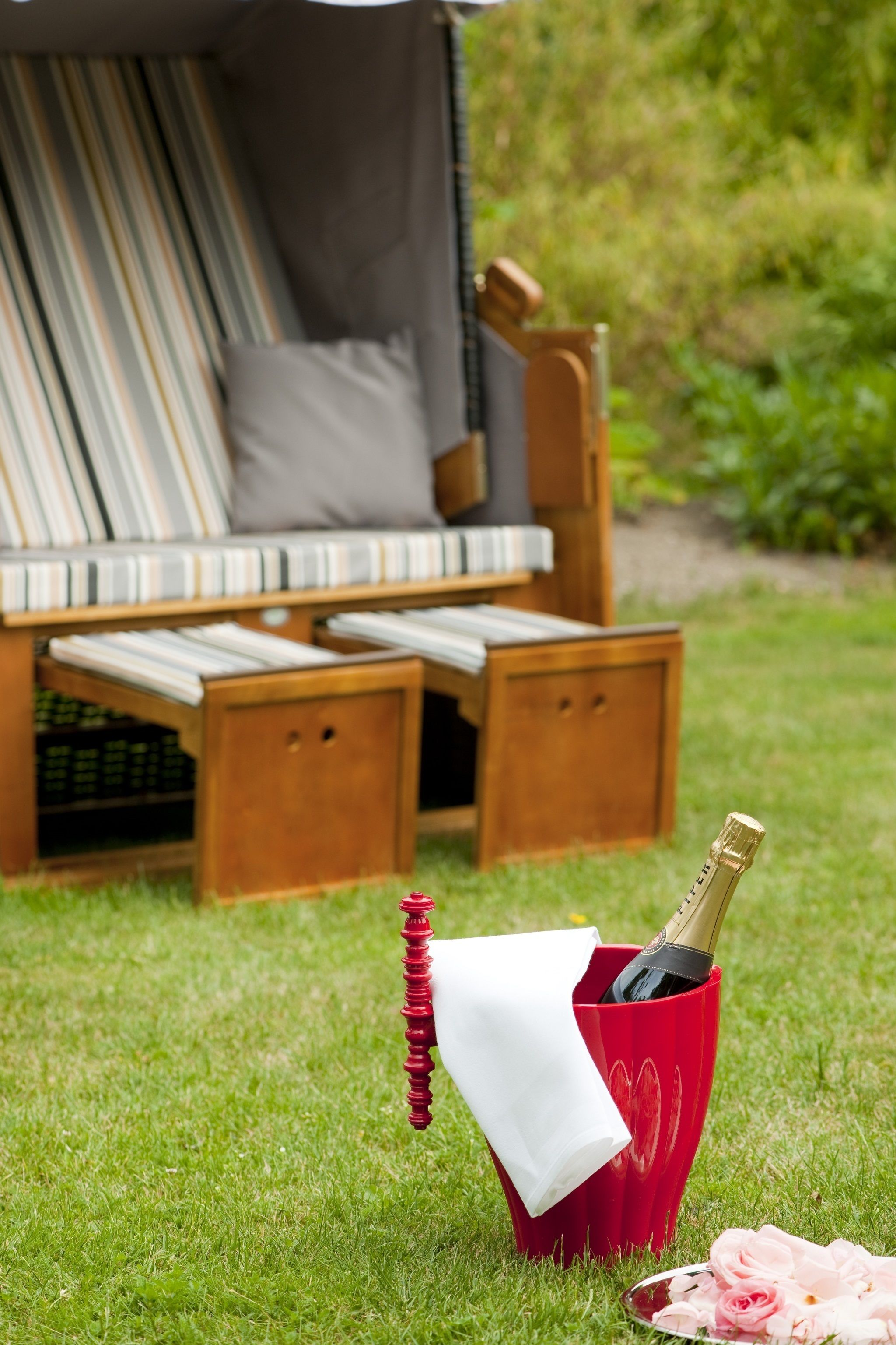 A wooden outdoor seating area with a red wine bottle and a white napkin in a heart-shaped holder on the grass.