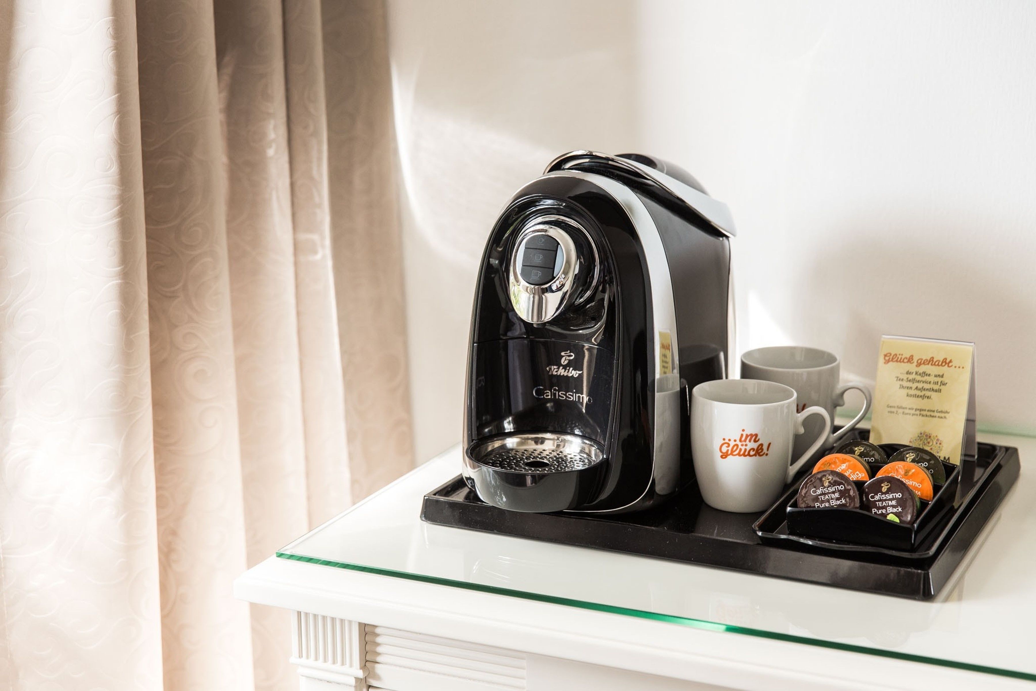 A coffee machine with a tray of coffee pods and two mugs on a glass table.