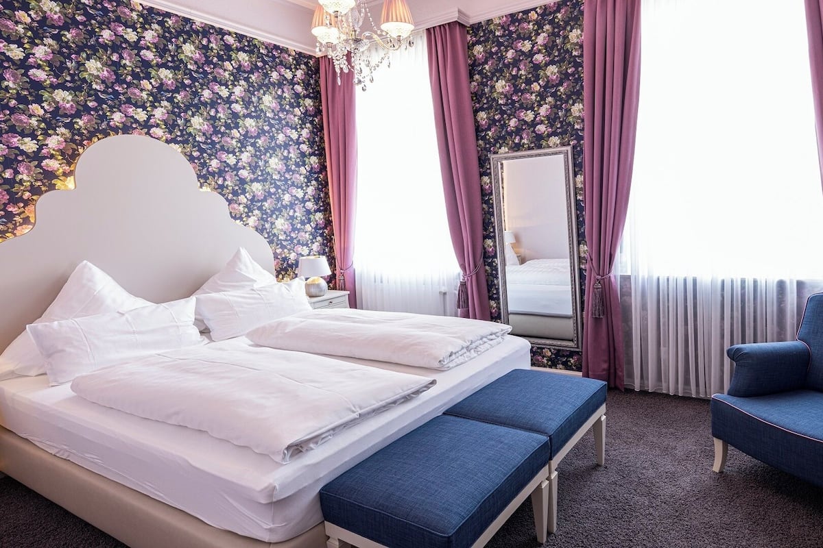 A hotel room with a large bed, a bench, a chair, a mirror, and floral wallpaper.