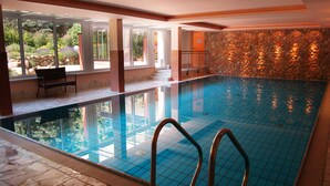 An indoor swimming pool with clear glass walls, a stone wall, and a bench by the window. - Hotel Heide Kröpke (Essel)